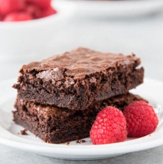 2 gluten free brownies sitting on white plate with 2 raspberries. gray background. more raspberries and brownies in background (at different angle)