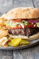 side angle of gray plate with 3 pickle slices, potato chips and gluten-free hamburger complete with toppings