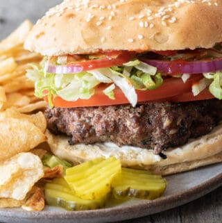 side angle of gray plate with 3 pickle slices, potato chips and gluten-free hamburger complete with toppings