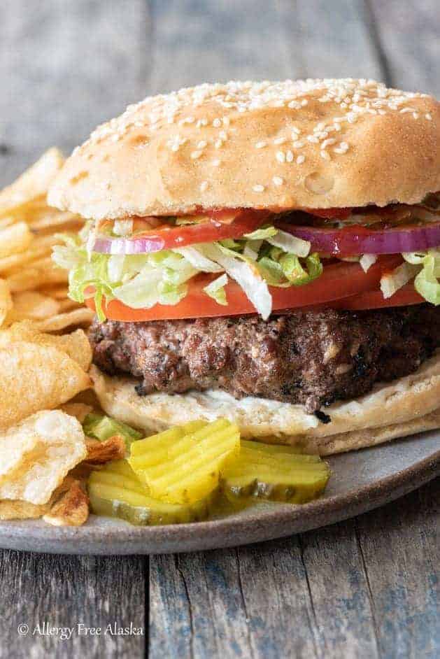 side angle of gray plate with 3 pickle slices, potato chips and gluten-free hamburger complete with toppings