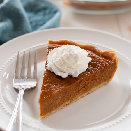 slice of gluten free pumpkin pie with dollop of whip cream on top. Fork sitting to side of pie, with blue napkin in background