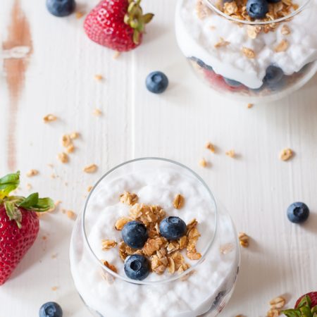 2 instant pot coconut milk yogurt parfaits sitting on white background, with berries and granola sprinkled on background (shot at different angle)