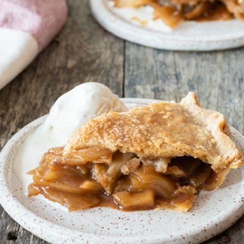 Slice of gluten-free apple pie sitting on white plate with scoop of ice cream. 2nd plate with pie in the far background.