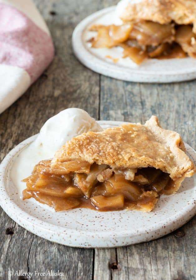 Slice of gluten-free apple pie sitting on white plate with scoop of ice cream. 2nd plate with pie in the far background.