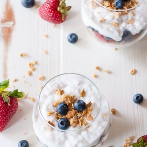 2 coconut milk parfaits in clear glasses with fresh berries and granola scattered across white background