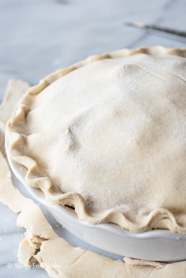 at different angle raw gluten-free apple pie sitting on marble pastry board with excess pie crust pieces