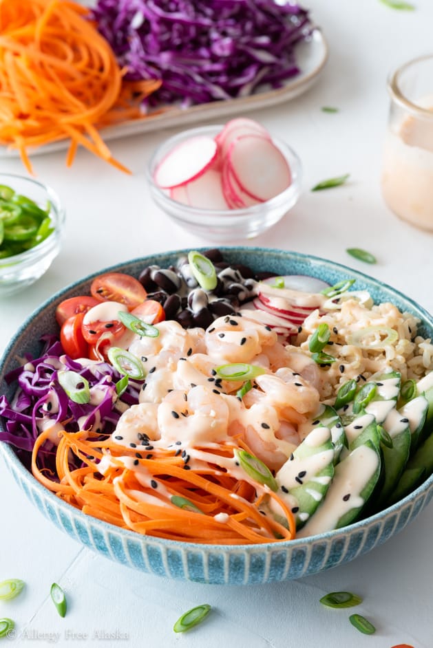 Side angle of shrimp poke bowl with Sriracha mayo on white background with sliced raddish, carrot ribbons, and sliced purple cabbage in background