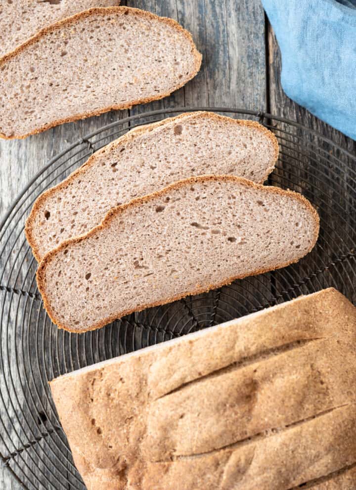 top down view of half sliced buckwheat bread sitting on wire cooling rack on gray/blue background