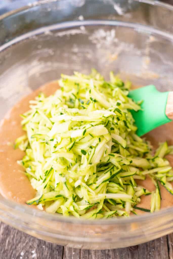 Raw zucchini muffin batter sitting in large clear bowl with a pile of grated zucchini