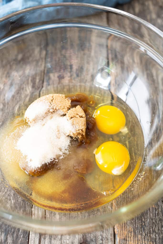 2 eggs, brown sugar, and white sugar in large clear glass bowl on wooden background