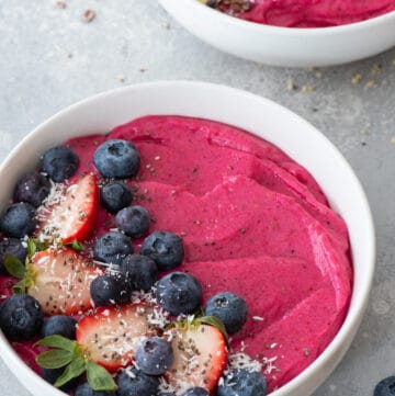 at different angle: vibrant pink dragon fruit smoothie in a white bowl, topped with blueberries, strawberries, and flaked coconut