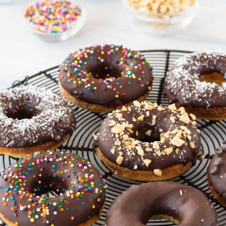 side angle view of chocolate glazed donuts sitting on black round cooling rack, with small bowls of sprinkles, chocolate chips, and peanuts in background