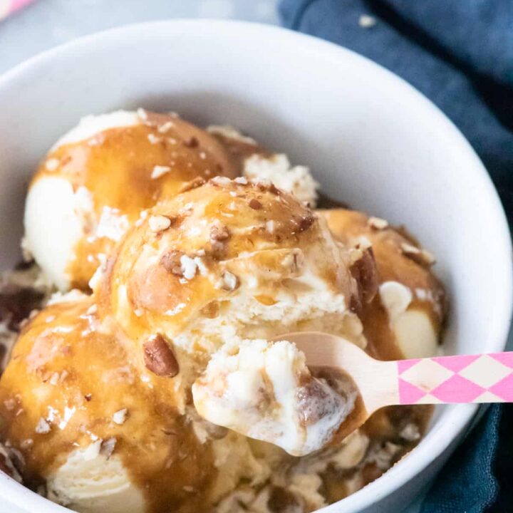 vanilla ice cream covered in vegan caramel sauce and chopped pecans, in white bowl on blue background. Pink and natural pattered spoon getting spoonful.