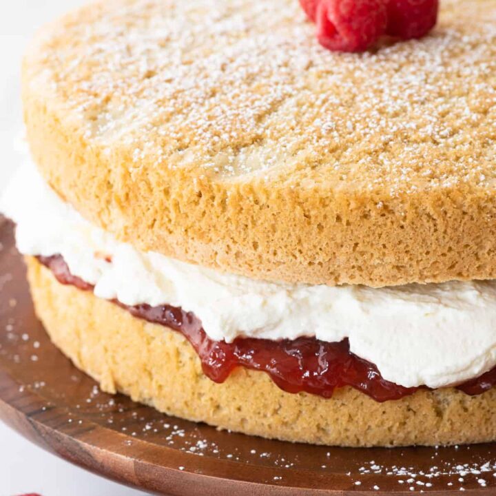 at different angle: vanilla gluten free cake rounds sandwiched with jam and whipped cream sitting on brown cake stand on white background