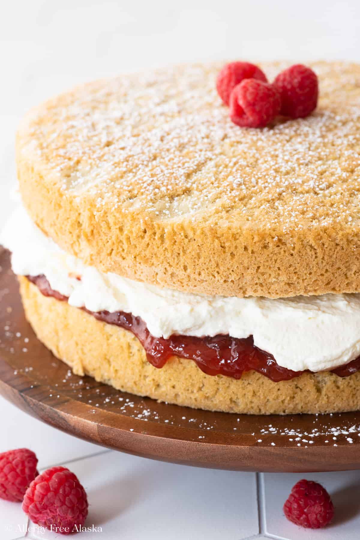 at different angle: vanilla gluten free cake rounds sandwiched with jam and whipped cream sitting on brown cake stand on white background