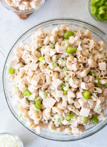 Top down view at different angle of gluten free macaroni salad in clear bowl on gray background. clear bowls of other ingredients in background