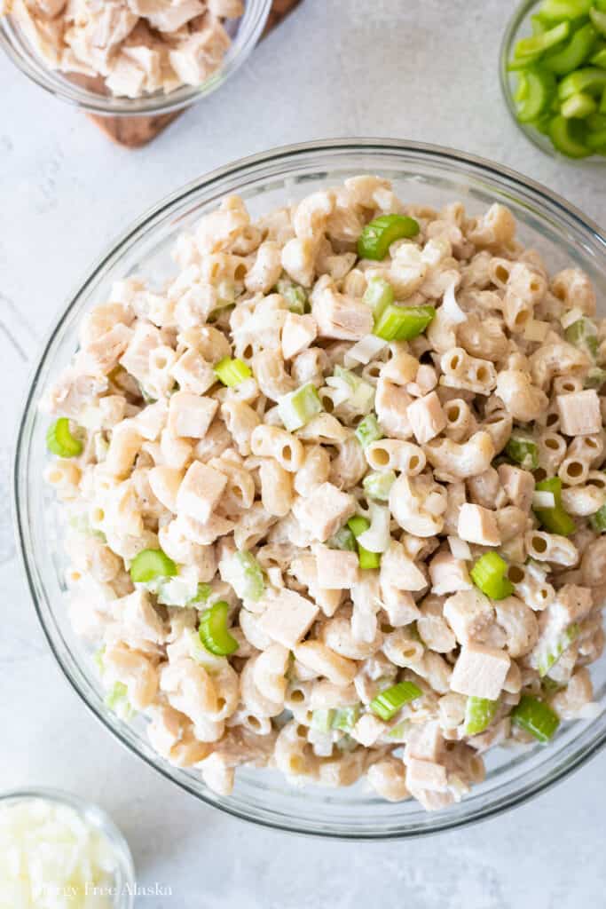 Top down view at different angle of gluten free macaroni salad in clear bowl on gray background. clear bowls of other ingredients in background